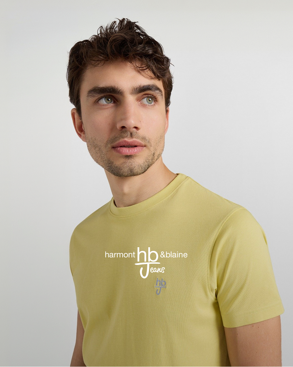 L'immagine ritrae il primo piano di un giovane uomo sorridente con capelli castani e mossi, orientato di trequarti verso sinistra mentre appoggia delicatamente il pugno alla guancia. Indossa una polo blu scuro con dettagli rosa sul colletto e sui bordi delle maniche, completata dalla scritta rosa "HBJ" sul colletto e dal logo sovrapposto "harmont & blaine hb Jeans" al centro.